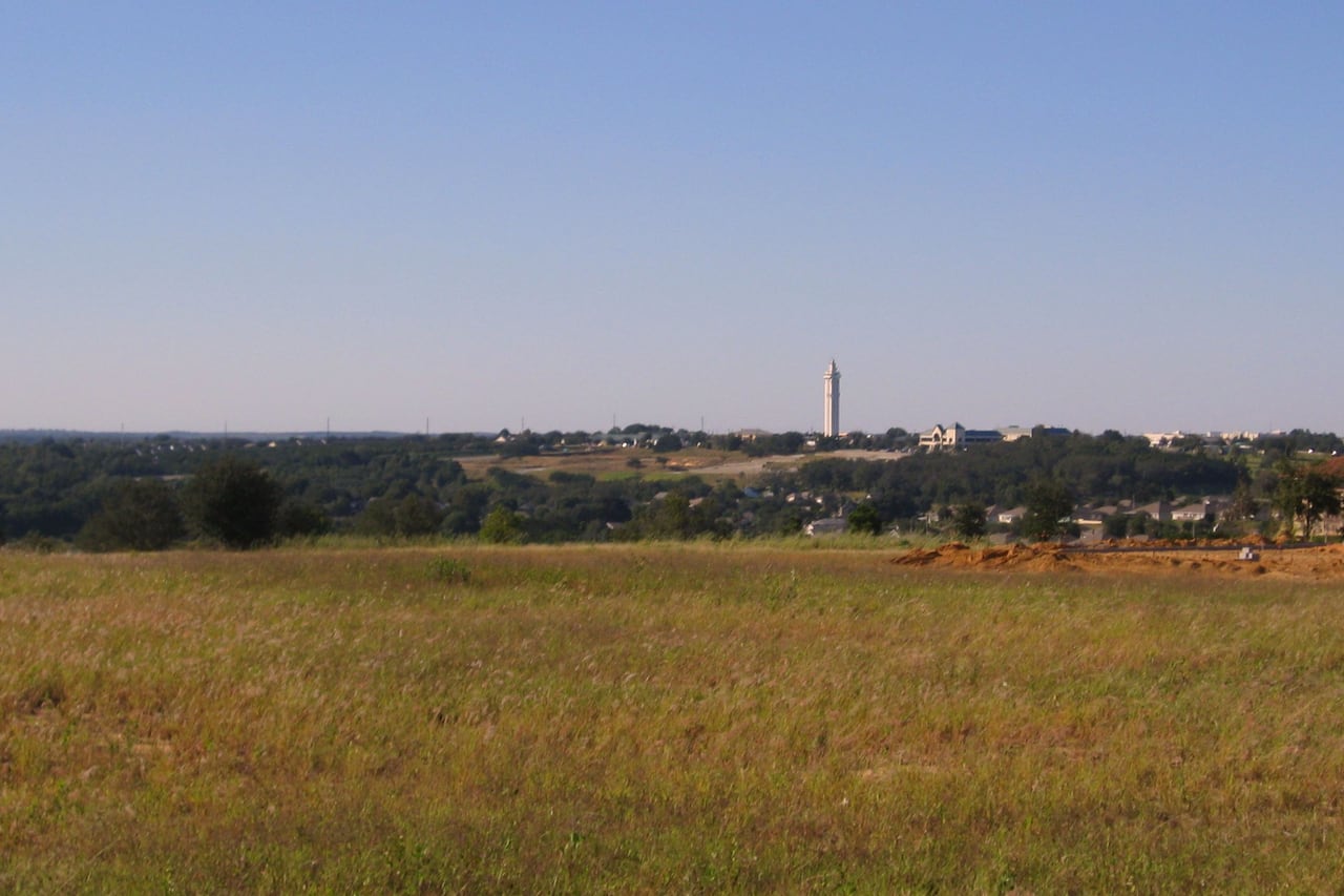 Campo aberto com morros arborizados ao fundo e uma torre fina no horizonte.
