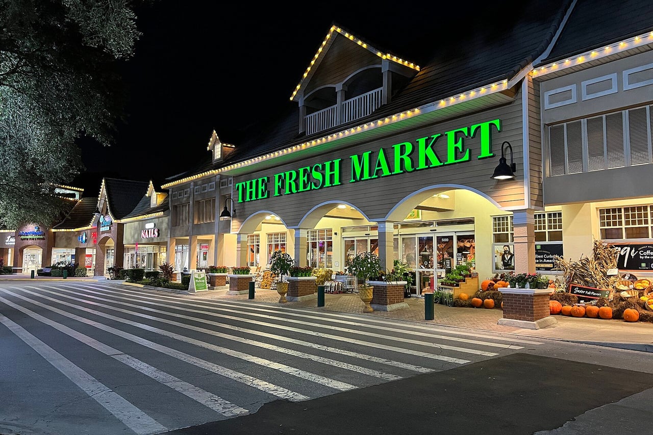 Fachada iluminada do supermercado The Fresh Market com faixa de pedestres na frente