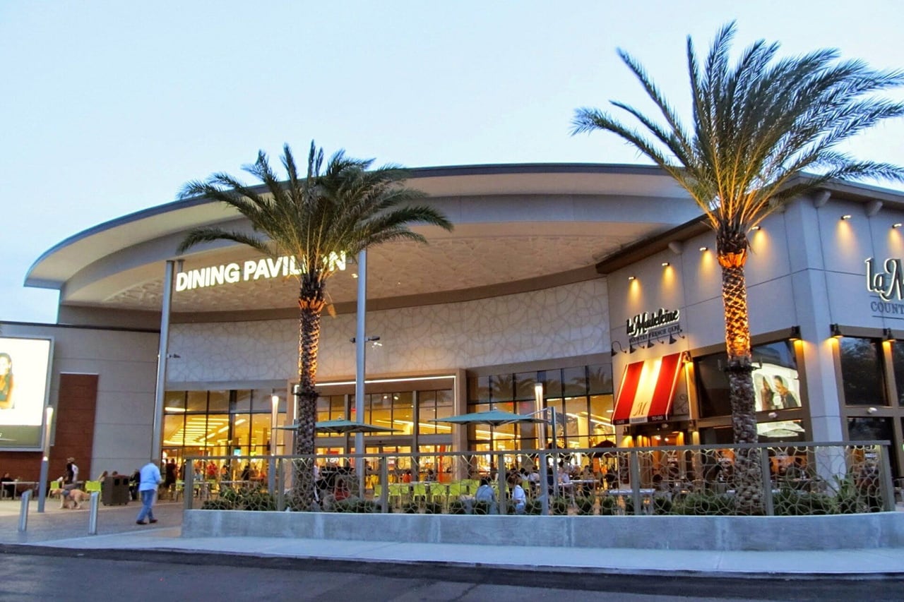 Entrada do Dining Pavilion do Florida Mall ao anoitecer, com duas palmeiras iluminadas e mesas ao ar livre