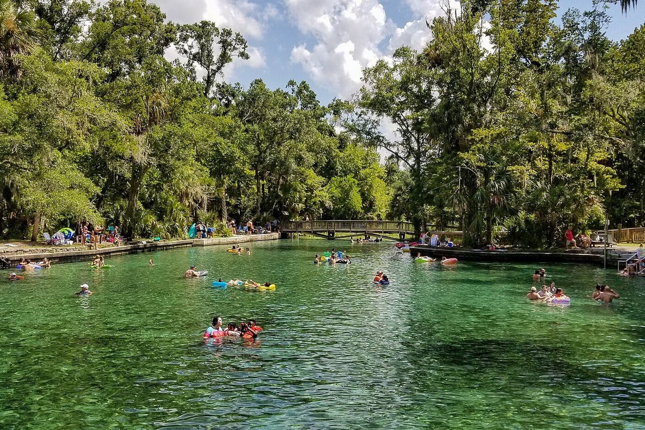 Piscina natural de água verde-clara cercada de mata, com pessoas boiando ao longe e uma passarela de madeira ao fundo