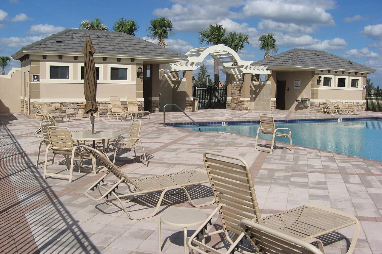 Deck de piscina com espreguiçadeiras, mesas e guarda-sol diante de um clube com pergolado