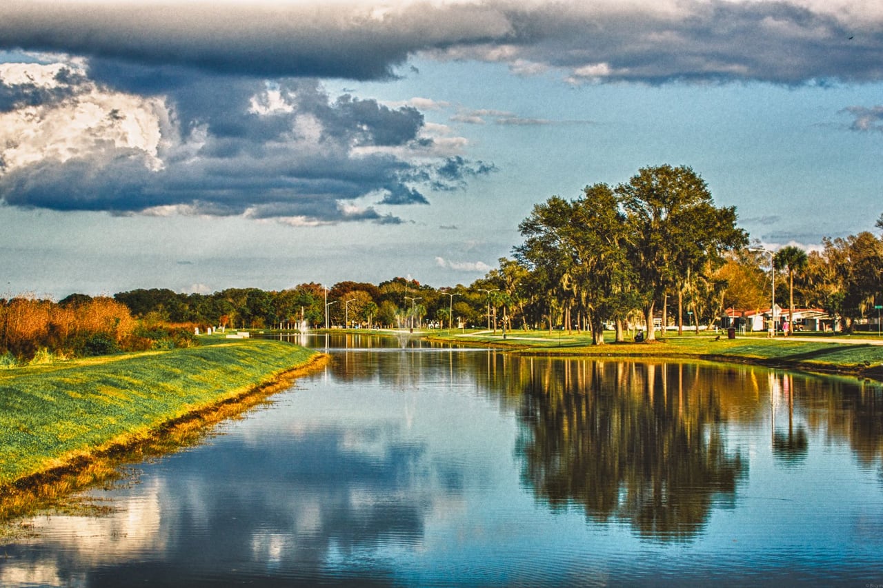 Canal calmo refletindo carvalhos e nuvens no gramado do Lakefront Park, em St. Cloud, no fim da tarde